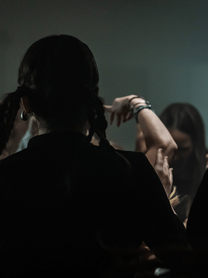 Frau mit geflochtenen Haaren und schwarzen Kleidungsstücken vor Spiegel in dunklem Raum