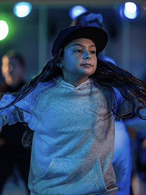 Mädchen mit grauem Hoodie und schwarzer Kappe tanzt in beleuchtetem Raum mit unscharfem Hintergrund