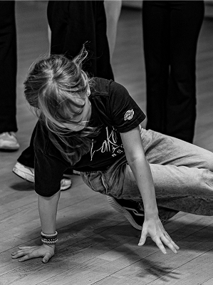 Schwarz-weiß-Foto eines Mädchens beim Breakdance auf Holzboden in Freizeitkleidung mit T-Shirt und Jeans