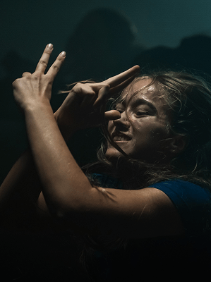 Junge Frau mit blonden Haaren in blauem Shirt macht im dunklen Raum mit Licht Schattenfiguren mit den Händen