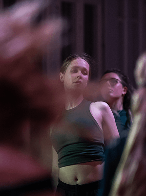 Junge Frau mit grünem Tanktop beim Tanz in gedämpft beleuchtetem Raum mit weiteren Personen im Hintergrund