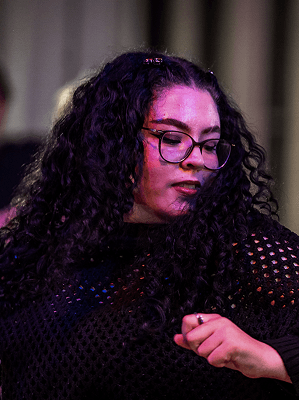 Frau mit langen lockigen dunklen Haaren und Brille in schwarzem Netzpullover bei Tanzbewegung vor dunklem Hintergrund