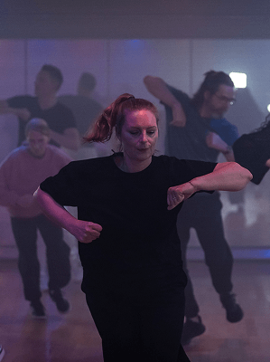 Frau im schwarzen T-Shirt tanzt konzentriert in einem Tanzstudio mit mehreren tanzenden Personen im Hintergrund