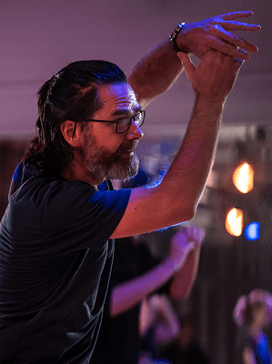 Mann mit Bart und Brille hebt beide Hände in einem Raum mit bunter Beleuchtung und unscharfem Hintergrund