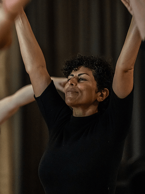 Frau mit kurzen lockigen Haaren in schwarzem Oberteil hebt Arme glücklich in Innenraum