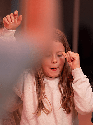 Mädchen mit langen braunen Haaren und rosa Pullover hebt Hände vor Augen in warm beleuchtetem Raum