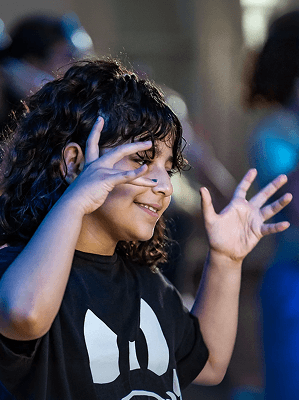 Kind mit lockigem Haar und schwarzem T-Shirt hebt Hände lachend während eines gesellschaftlichen Events