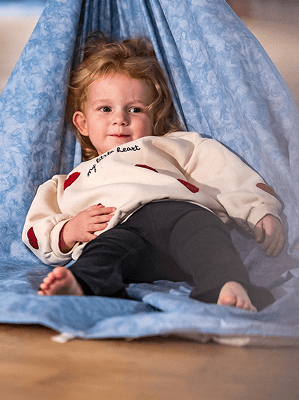 Kleines Mädchen liegt entspannt in einem hellblauen Stoffzelt auf Holzboden trägt hellen Pullover und dunkle Leggings