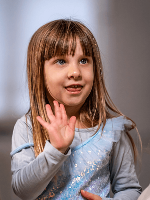 Junges Mädchen mit braunen Haaren und blau-grauem Pullover hebt die Hand in heller Innenraumumgebung