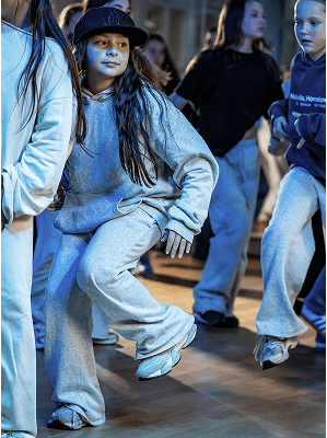 Mädchen in grauem Hoodie und Jogginghose mit schwarzer Cap tanzt in Tanzstudio mit anderen Jugendlichen