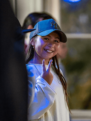 Junges Mädchen mit blauer Baseballkappe und weißem Pullover lächelt und zeigt ein Peace-Zeichen in urbaner Umgebung bei Abendlicht