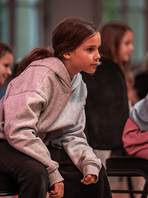 Mädchen mit grauem Kapuzenpullover sitzt gespannt auf Stuhl in Innenraum mit weiteren Kindern im Hintergrund