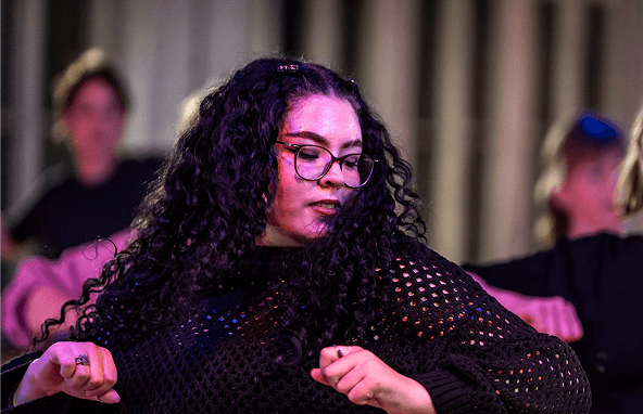 Junge Frau mit langen lockigen Haaren und Brille tanzt in schwarzem Netzpullover vor verschwommenem Hintergrund