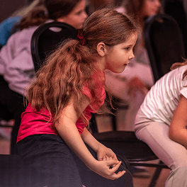 Mädchen mit roten Haaren und rotem Shirt konzentriert sich bei einem Gruppenunterricht auf schwarzen Stühlen