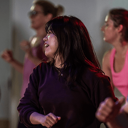 Frau mit dunklen Haaren und Brille beim Tanzkurs in Sportkleidung in einem Fitnessraum mit anderen Teilnehmerinnen