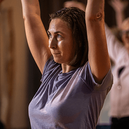 Frau in lila T-Shirt hebt im Fitnesskurs konzentriert die Arme über den Kopf vor unscharfem Hintergrund