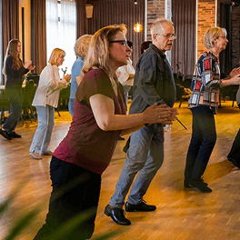 Gruppe von Erwachsenen beim Tanzkurs in einem hellen Raum mit Holzboden und Backsteinwänden