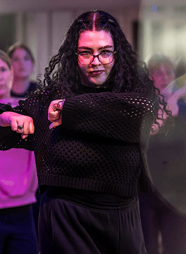 Frau mit lockigem Haar und Brille tanzt in schwarzem Netzpullover in einer Tanzgruppe in Innenraum