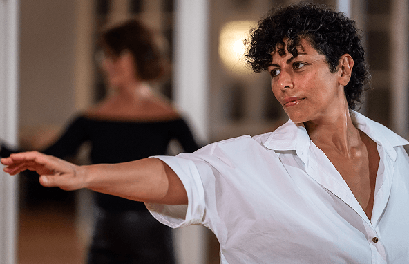 Frau mit lockigem Haar trägt weiße Bluse und macht Dehnübung im Innenraum mit unscharfer Person im Hintergrund