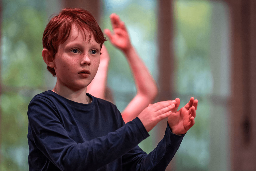 Junge mit roten Haaren klatscht konzentriert im Tanzunterricht, im Hintergrund weitere Kinder in Bewegung