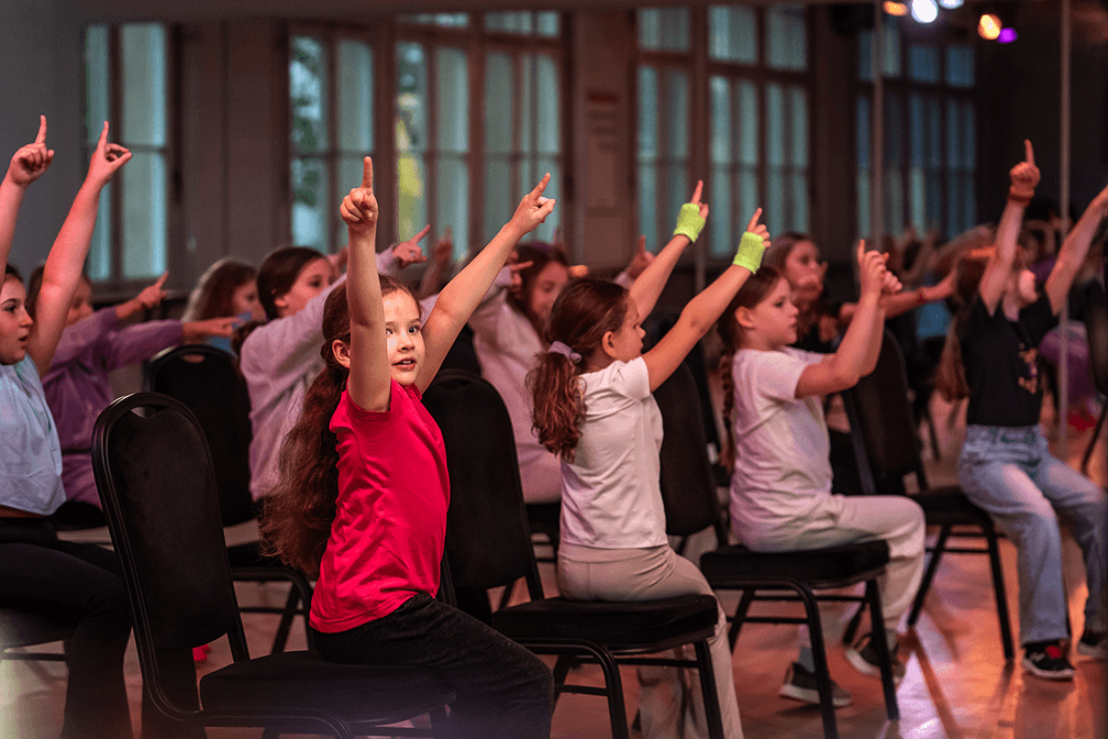 Kinder sitzen auf Stühlen im Tanzsaal und strecken synchron die Arme nach oben während einer choreografierten Übung