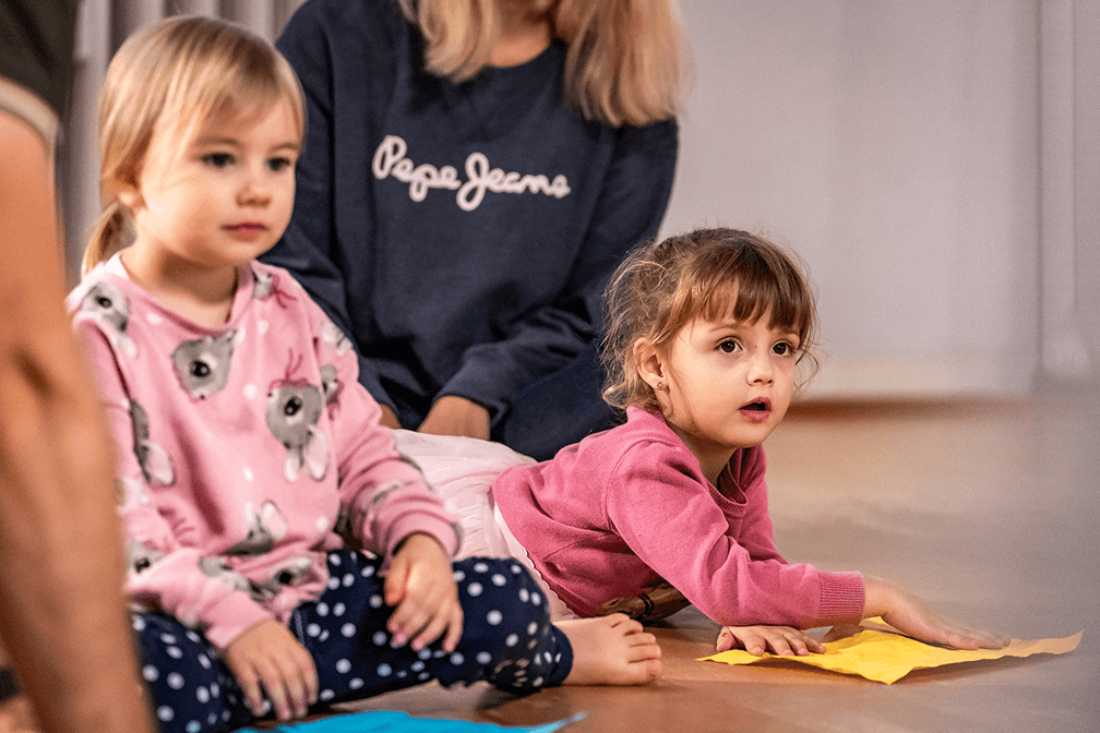 Zwei Kleinkinder sitzen auf dem Boden im Tanzsaal und erkunden bunte Tücher während des Kindertanzunterrichts, im Hintergrund eine erwachsene Begleitperson