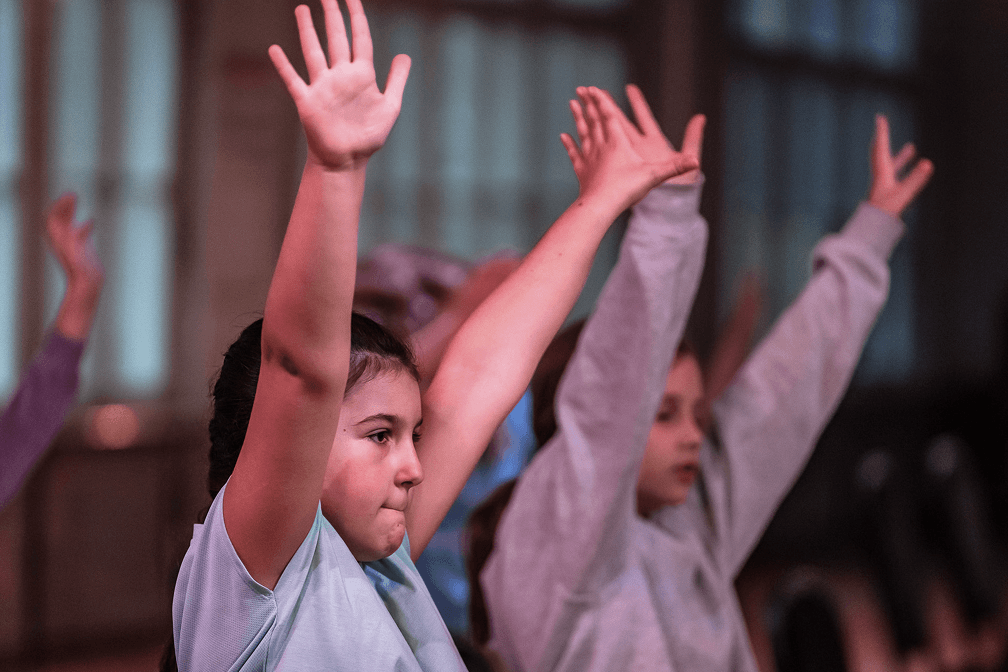 Mehrere Kinder stehen im Tanzsaal und strecken die Arme nach oben, während sie gemeinsam eine Tanzbewegung ausführen