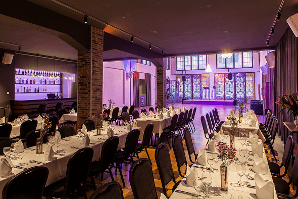 Elegantes Restaurant mit lang gedeckten Tischen, schwarzen Stühlen, beleuchtetem Barbereich und Tanzfläche mit großen Fenstern