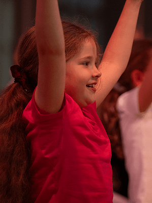 Mädchen mit roten Haaren und rotem T-Shirt hebt freudig beide Arme in einer hell beleuchteten Umgebung
