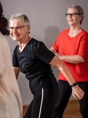 Ältere Frau in schwarzem Sportdress macht Dehnübungen im Fitnessstudio mit weiteren Teilnehmerinnen im Hintergrund
