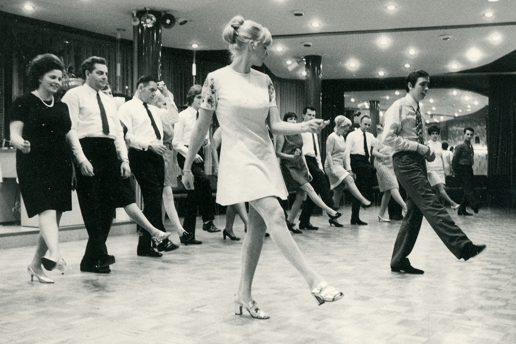 Schwarzweißaufnahme von Tanzgruppe mit Männern und Frauen in 60er-Jahre-Kleidung bei Tanzstunde in Saal mit Spiegelwänden