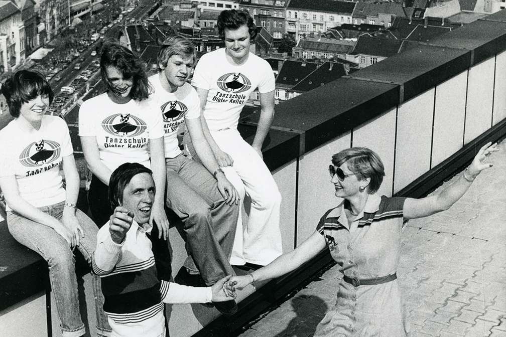 Schwarzweißfoto einer Tanzgruppe auf einem Dach mit T-Shirts der Tanzschule Dieter Keller vor Stadtkulisse