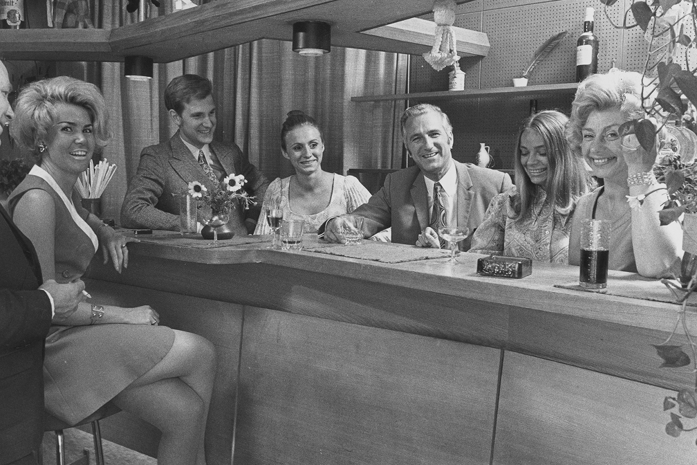 Schwarzweißfoto einer Gruppe elegant gekleideter Menschen am Holztresen in Bar mit Getränken und Dekoration aus den 1970er Jahren