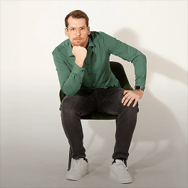 Junger Mann mit Brille in grühem Hemd und schwarzen Jeans sitzt nachdenklich auf einem grünen Stuhl vor hellem Hintergrund
