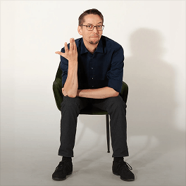 Mann mit Brille sitzt auf grünem Stuhl vor weißem Hintergrund und gestikuliert mit linker Hand